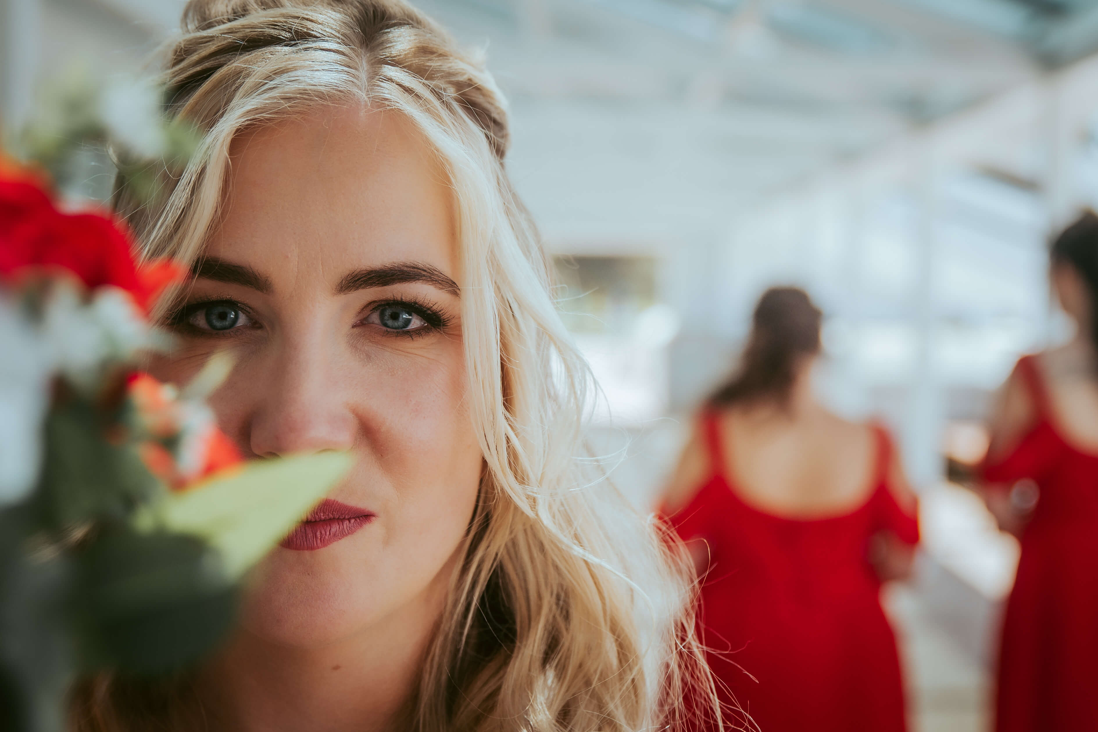 Close up portrait image of women with blonde hair and minimal makeup, while the background is blurred with women standing in red dresses.